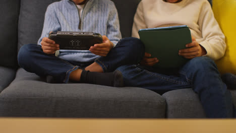 Close-Up-Of-Two-Young-Boys-Sitting-On-Sofa-At-Home-Playing-Games-Or-Streaming-Onto-Digital-Tablet-And-Handheld-Gaming-Device-1